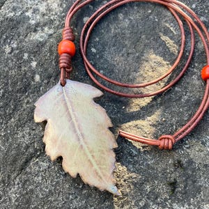 May include: A leaf-shaped pendant necklace with a brown leather cord. The pendant is a light beige color with a detailed leaf vein pattern. Two orange beads accent the cord. The necklace is displayed on a gray stone surface.