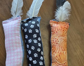 May include: Three cat toys with fabric bodies and feathers. One toy has a pink and white houndstooth pattern, another has a black background with white paw prints, and the third has an orange and red floral design. Each toy has a denim border.