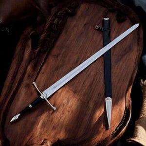 May include: A silver-toned steel sword with a black grip and a cross-shaped guard, accompanied by a matching black scabbard featuring silver-toned metal accents at the tip and throat, displayed together on a dark, polished wooden surface.