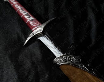 May include: A dagger featuring a red wood-grain handle with silver accents, a silver crossguard with intricate engravings, and a silver blade with etched script. The dagger is partially inserted into a brown sheath with ornate silver metal trim.