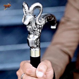 May include: Silver-coloured walking cane with a ram's head handle. The handle has intricate detailing and a brass collar where it meets the black wooden shaft. The cane is held by a person.
