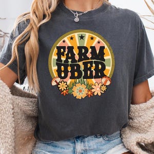 May include: A dark gray t-shirt with a retro-style graphic that says "Farm Uber" in a circle with a yellow sunburst and flowers.