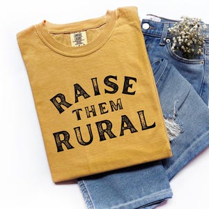 Könnte beinhalten: Senfgelbes T-Shirt mit dem schwarzen Text "RAISE THEM RURAL", das auf einer blauen Jeans liegt. Die Jeans hat einen Used-Look und einen kleinen Strauß weißer Blumen in der Tasche.