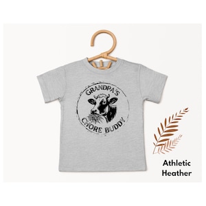 Könnte beinhalten: Graues T-Shirt mit kurzen Ärmeln und einem schwarz-weißen Grafikdruck einer Kuh, die Heu frisst. Die Grafik befindet sich in einem Kreis mit dem Text "Grandpa's Chore Buddy".