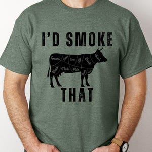Könnte beinhalten: Grünes T-Shirt mit dem Text "I'D SMOKE THAT" über einer schwarzen Abbildung einer Kuh mit beschrifteten Fleischstücken. Ein lässiges Kleidungsstück.