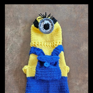 May include: A handmade crochet pet costume resembling a Minion character. The costume features a yellow body with blue overalls, a single eye, and black hair. The outfit is designed to fit a small dog or cat.