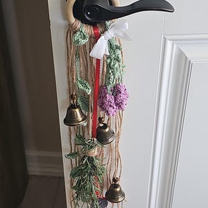 Może przedstawiać: Ozdoba na drzwi z napisem "Witches Bells". Posiada drewniany pierścień, sznurek jutowy, szydełkowe zielone liście i fioletowe kwiaty, czerwoną wstążkę, białą kokardę i trzy brązowe dzwonki. Zawieszka jest przymocowana do czarnej klamki.