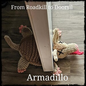 May include: A crocheted armadillo doorstop in brown and pink with a pink tongue sticking out. The armadillo is lying on its back with its legs in the air. The text "From Roadkill to Doorsil" is at the top of the image and "Armadillo" is at the bottom.