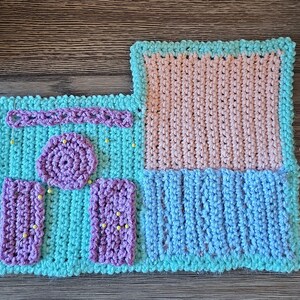 May include: A handmade crochet project in shades of blue, pink, and purple. The piece features a rectangular design with various textured sections, including a circular element and two rectangular pieces. Yellow pins are placed throughout the project.