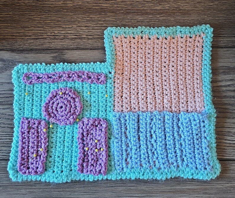 May include: A handmade crochet project in shades of blue, pink, and purple. The piece features a rectangular design with various textured sections, including a circular element and two rectangular pieces. Yellow pins are placed throughout the project.