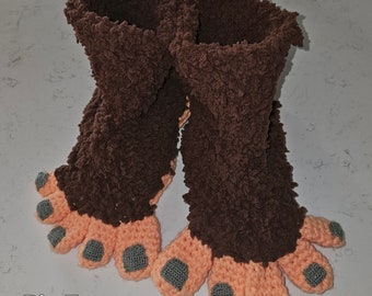 May include: A pair of brown fuzzy slippers shaped like Bigfoot feet. The toes are crocheted in a peach color with gray toenails.