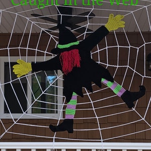 May include: A crocheted witch figure with a black hat, green and purple striped legs, and yellow hands is caught in a white spider web. The text "Caught in the Web" is written in green letters above the witch.
