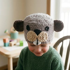 May include: A handmade crochet beaver hat. The hat is mainly grey with brown ears and a beige muzzle. Black button eyes and a white and black nose complete the beaver face. The hat is worn by a child.