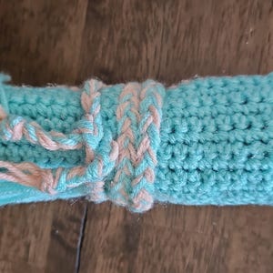 May include: A hand-crocheted item in a rolled-up shape, primarily turquoise blue with a braided band and tassels in a light peach color. The item is made of yarn and appears soft and textured.