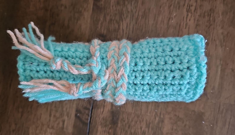 May include: A hand-crocheted item in a rolled-up shape, primarily turquoise blue with a braided band and tassels in a light peach color. The item is made of yarn and appears soft and textured.