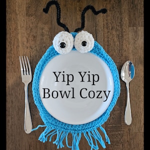 May include: A blue crocheted bowl cosy with fringe, featuring two white googly eyes with black pupils and two black pipe cleaner antennae. The text "Yip Yip Bowl Cozy" is written in black on a white bowl inside the cosy.
