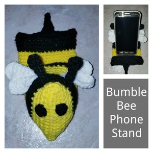 Könnte beinhalten: Ein gehäkelter Hummel-Handyständer. Die Hummel ist gelb und schwarz mit weißen Flügeln. Der Handyständer ist so konzipiert, dass er ein Smartphone aufrecht hält.