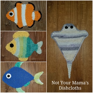 May include: Four crocheted dishcloths in the shape of sea creatures: a blue fish, a green and yellow fish, an orange and white clownfish, and a gray stingray. The dishcloths are displayed on a wooden background with the text "Not Your Mama's Dishcloths".