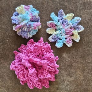 May include: Three crocheted floral appliques. One is a solid bright pink flower with layered petals. The other two are multi-colored flowers featuring a blend of pastel yellow, light blue, and lavender yarn in a variegated pattern with distinct petal shapes.