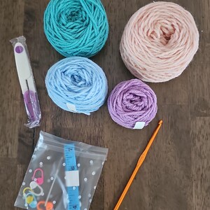 May include: A craft kit with four balls of yarn in teal, light pink, light blue, and purple. The kit also includes a crochet hook, a seam ripper, stitch markers, and a blue measuring tape in a clear bag.