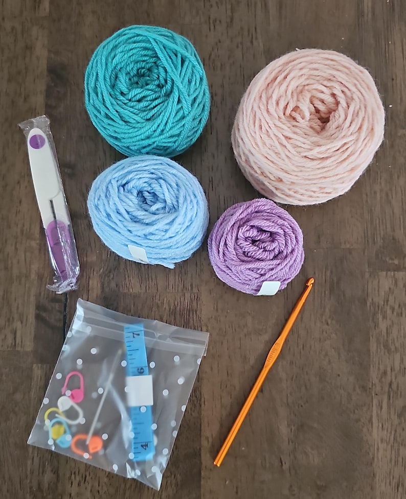 May include: A craft kit with four balls of yarn in teal, light pink, light blue, and purple. The kit also includes a crochet hook, a seam ripper, stitch markers, and a blue measuring tape in a clear bag.