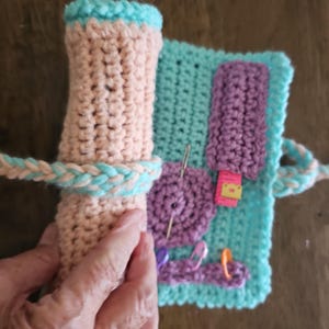 May include: A handmade crochet needle case in shades of peach, teal, and purple. The case is rolled up and secured with a braided teal and peach strap. The open case reveals a teal background with purple crochet details and sewing pins.
