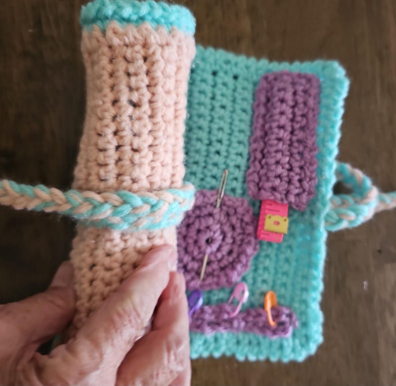 May include: A handmade crochet needle case in shades of peach, teal, and purple. The case is rolled up and secured with a braided teal and peach strap. The open case reveals a teal background with purple crochet details and sewing pins.