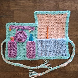 May include: Crocheted organizer in shades of blue, purple, and beige, featuring pockets and loops for crochet tools. Includes a braided strap and tassel details. The organizer holds a crochet hook, stitch markers, and other accessories.