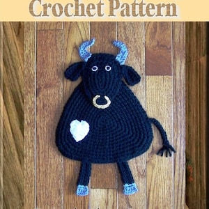 Puede incluir: Un posavasos de crochet negro en forma de toro con cuernos azules, un anillo de nariz dorado y un corazón blanco en un lado. El posavasos está sobre una superficie de madera. Designs by: Sharon Santorum.