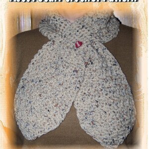 May include: A beige crochet scarf with a textured pattern, tied in a knot around the neck. The scarf has a small red button in the centre. The text "Ascot Scarf Crochet Pattern" is at the top of the image and "Design by Sharon Santorum" is at the bottom.