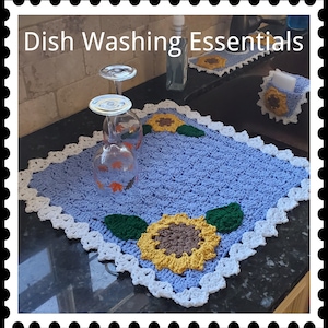 May include: A blue crocheted dish drying mat with a white crocheted border and a sunflower design. The mat is on a black countertop with two glasses and a bottle of hand soap.