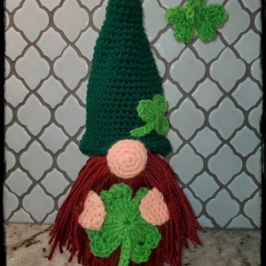 May include: A green crocheted gnome with a red beard and a shamrock on its hat. The gnome is holding a green shamrock in its hands. The gnome is sitting on a white tiled surface. The text "St. Patrick's Day Wine Buddy" is written below the gnome.