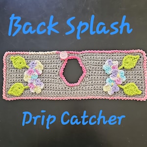 May include: A crocheted grey drip catcher with pink trim, a pink button, and a central opening. It features floral and leaf accents in pastel colours and the text "Back Splash Drip Catcher" in blue.