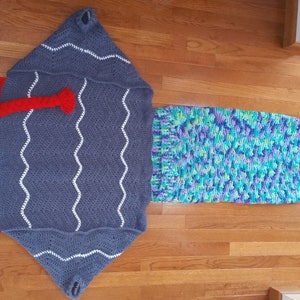 Puede incluir: Un saco de dormir de cola de sirena de crochet con una cola azul, verde y morada y una parte superior con capucha gris y roja.