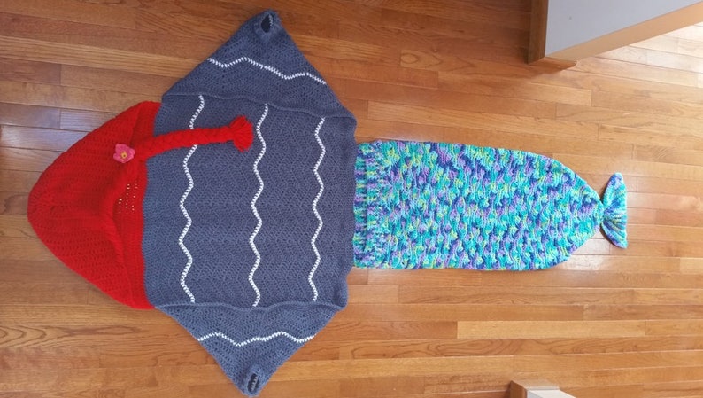 Puede incluir: Un saco de dormir de cola de sirena de crochet con una cola azul, verde y morada y una parte superior con capucha gris y roja.