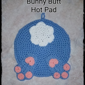 Könnte beinhalten: Ein handgefertigter, blauer Topflappen aus Häkelarbeit in Form eines Hasen-Hinterns, mit einem weißen Schwanz und rosa Pfoten. Der Text "Bunny Butt Hot Pad" ist oben zu sehen. Dieses skurrile Küchenaccessoire sorgt für Spaß.