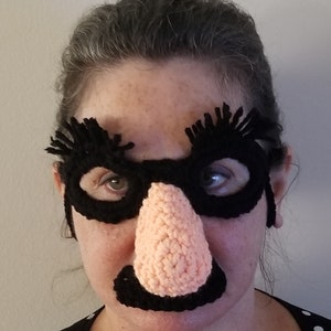 May include: A person wearing a black crocheted mask with a large pink crocheted nose and black mustache. The mask has eye holes and long black eyelashes.
