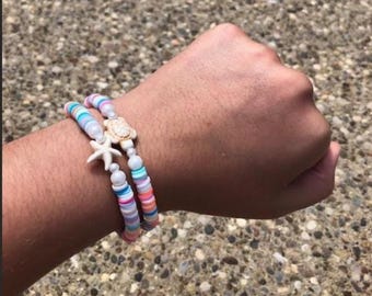 Puede incluir: Dos pulseras de cuentas con temática playera. Una pulsera presenta un dije de estrella de mar, la otra un dije de tortuga. Las pulseras tienen cuentas planas y coloridas en tonos rosa, azul y blanco. Las pulseras se llevan en la muñeca.