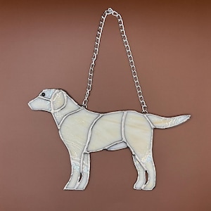May include: A stained glass hanging of a white labrador retriever. The dog is standing with its head turned to the left. The glass is iridescent and has a chain for hanging.