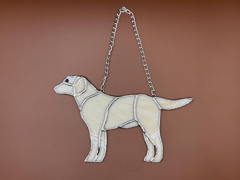 May include: A stained glass hanging of a white labrador retriever. The dog is standing with its head turned to the left. The glass is iridescent and has a chain for hanging.
