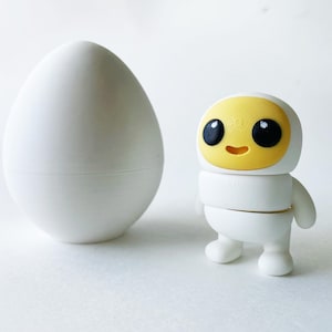 Puede incluir: Un juguete blanco con forma de huevo con un juguete blanco más pequeño con forma de huevo en su interior. Un robot de juguete blanco con una cara amarilla y ojos negros se encuentra junto a los huevos.