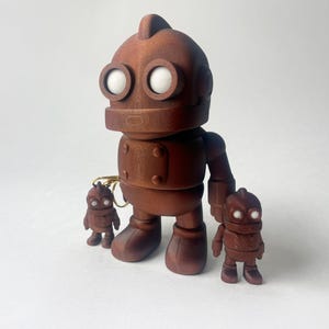 May include: A wooden robot figure with two smaller robot figures. The robots are brown with white eye details. The large robot has a key chain attached. The figures are standing on a white surface.