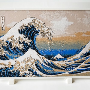 May include: Framed artwork depicting the iconic Japanese woodblock print, "The Great Wave off Kanagawa." The image features a large, stylized wave in shades of blue and white, with Mount Fuji in the background. The artwork is in a white frame.