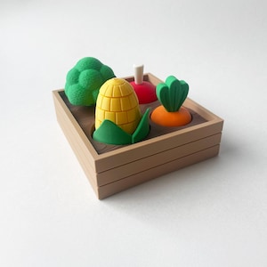 Puede incluir: Una caja de madera llena de verduras de juguete, incluyendo un brócoli verde, una mazorca de maíz amarilla, un tomate rojo y una zanahoria naranja.