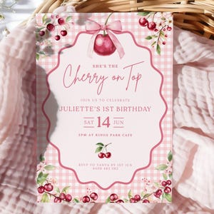 Op de afbeelding: Een roze en witte ruitjes verjaardagsuitnodiging met de tekst "She's the Cherry on Top". De uitnodiging is voor Juliette's 1e verjaardag, op 14 juni. Kersen en bloemaccenten versieren de rand.