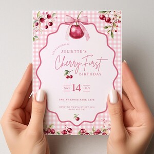 Op de afbeelding: Een roze en witte uitnodigingskaart met een kersenthema. De kaart leest "Cherry First Birthday" en bevat de datum "SAT 14 JUN". De kaart wordt door twee handen vastgehouden.