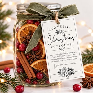 May include: A clear glass jar filled with dried orange slices, star anise, cinnamon sticks, and red cranberries. An olive green ribbon is tied around the lid, holding a white tag that reads: STOVETOP Christmas POTPOURRI. Instructions included.