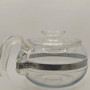 Könnte beinhalten: Eine klare Glasteekanne mit rundem Körper und gebogenem Griff. Ein silberfarbenes Metallband umschließt die Mitte der Teekanne. Der Deckel ist ebenfalls aus klarem Glas. Die Teekanne ist zum Aufbrühen von Tee bestimmt.