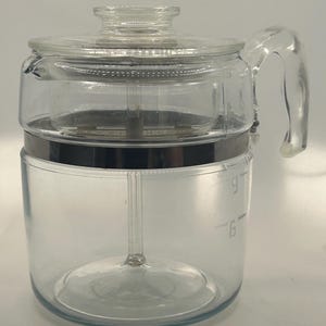 May include: A clear glass coffee percolator with a glass lid and handle. The percolator has a metal band and a central glass tube. Measurement markings for 6 and 9 cups are visible on the side.