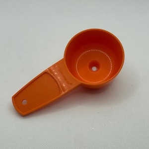May include: An orange plastic measuring scoop with a round bowl and a handle. The handle has a small hole at the end. The scoop is made of a durable plastic material and is designed for measuring dry or liquid ingredients.